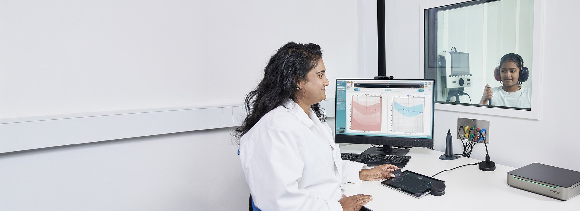 A girl is sitting in a sound booth undergoing a hearing test conducted by an audiologist who is operating the Touch Keyboard of the Equinox Evo.