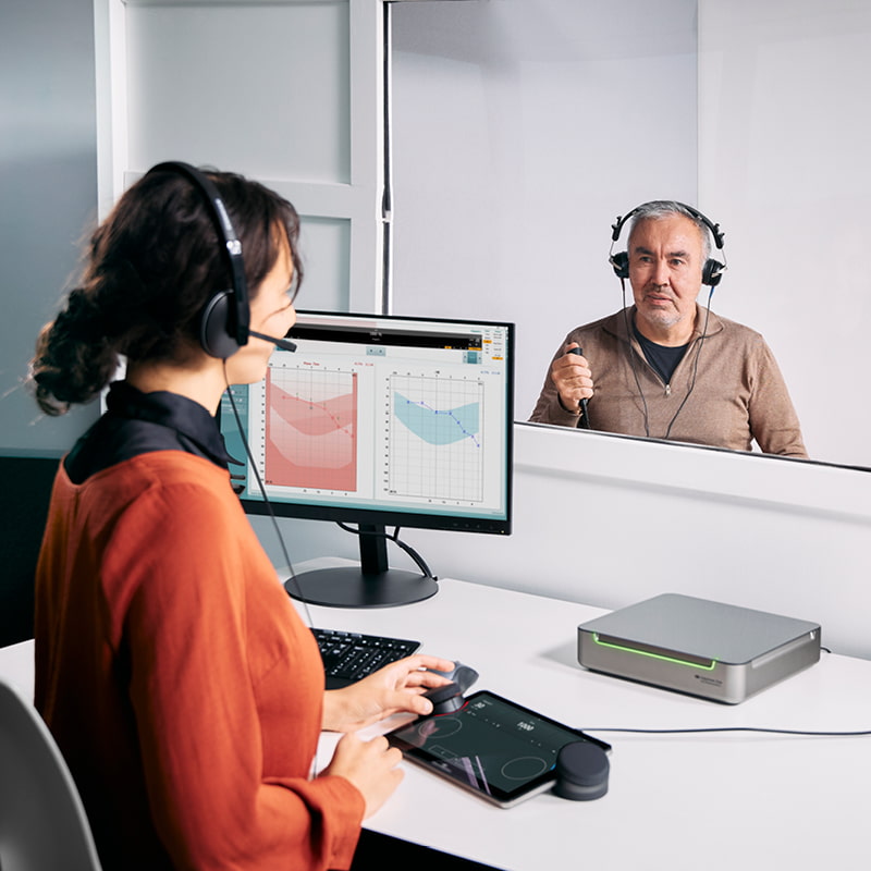 Egy idős férfi audiometriai vizsgálaton vesz részt, miközben az audiológus az érintőképernyős billentyűzetet használja