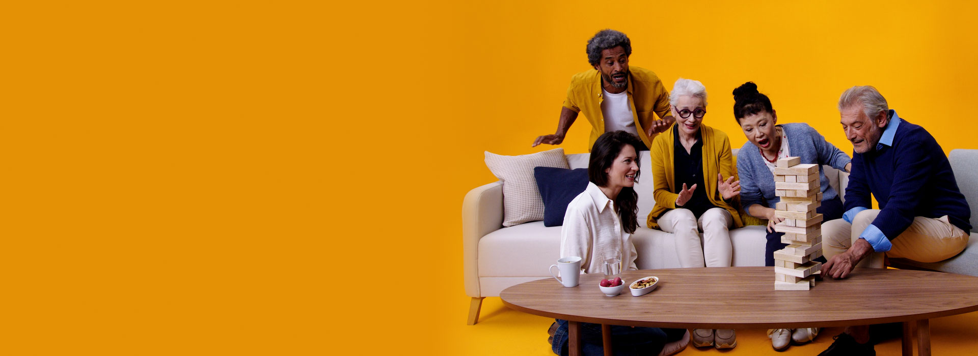A group of five adults and seniors, including a man and woman wearing Philips HearLink hearing aids sit on a couch playing Jenga together on a large coffee table