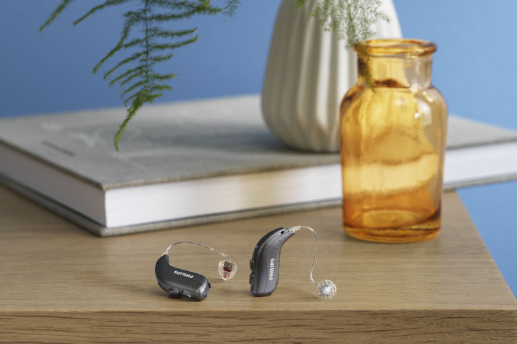 Philips HearLink non-rechargeable hearing aids placed on a wooden table, with a magazine, glasses and yellow plant pot