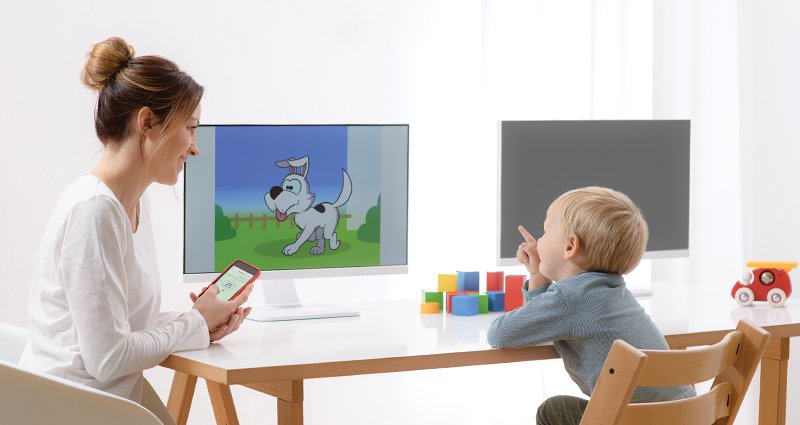 A clinician is seated at  a table with a child testing his hearing using the MedRx VRA system. The clinician holds an ipod she uses to record his responses. Rewards are displayed on the monitors in front of the child. 