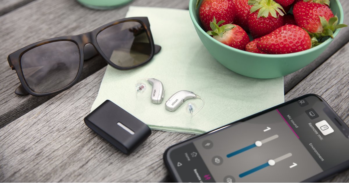 Des aides auditives Oticon posées sur un banc à coté d'un smartphone ouvert sur l'application Oticon ON , un bol de fraises et une paire de lunette