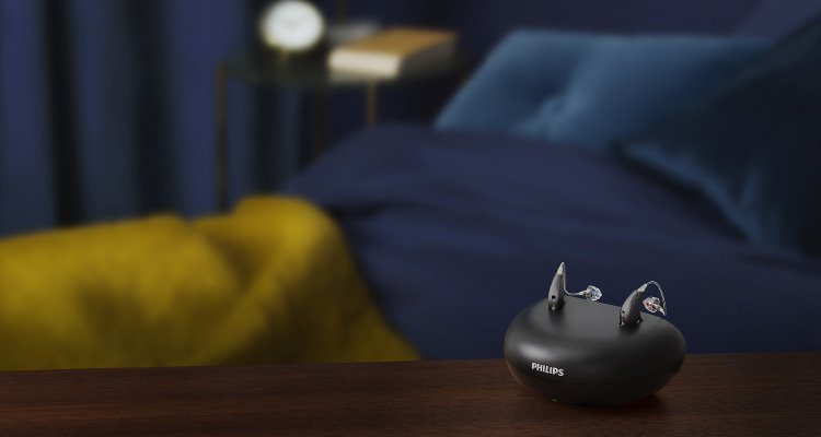 Τα επαναφορτιζόμενα ακουστικά βοηθήματα Philips HearLink miniRITE T R τοποθετημένα στον φορτιστή τους, επάνω στο τραπεζάκι στης κρεβατοκάμαρας. 