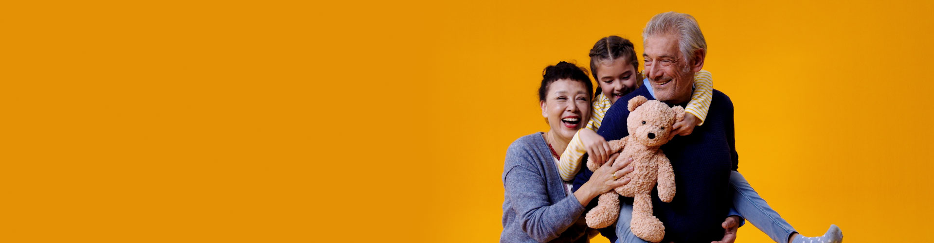 A granddaughter holds a teddy bear while sitting on the back of her grandfather who wears Philips HearLink hearing aids. Her mother also reaches out to hold her daughter.