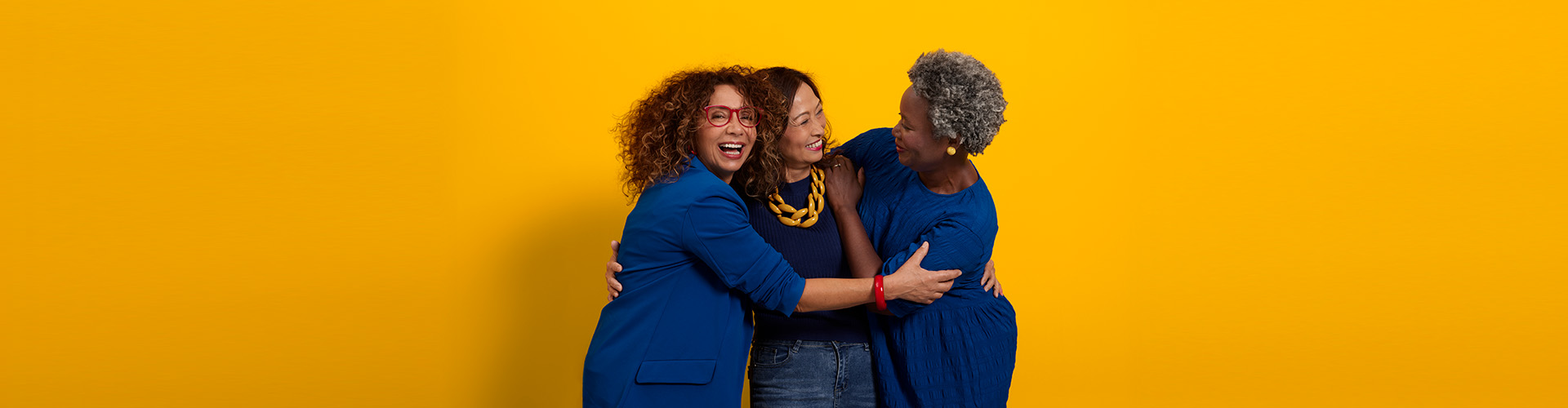 Three middle-aged women of different ethnicities dressed in blue hugging and smiling.