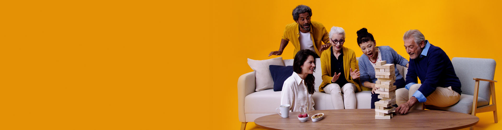 A group of five adults and seniors, including a man and woman wearing Philips HearLink hearing aids sit on a couch playing Jenga together on a large coffee table