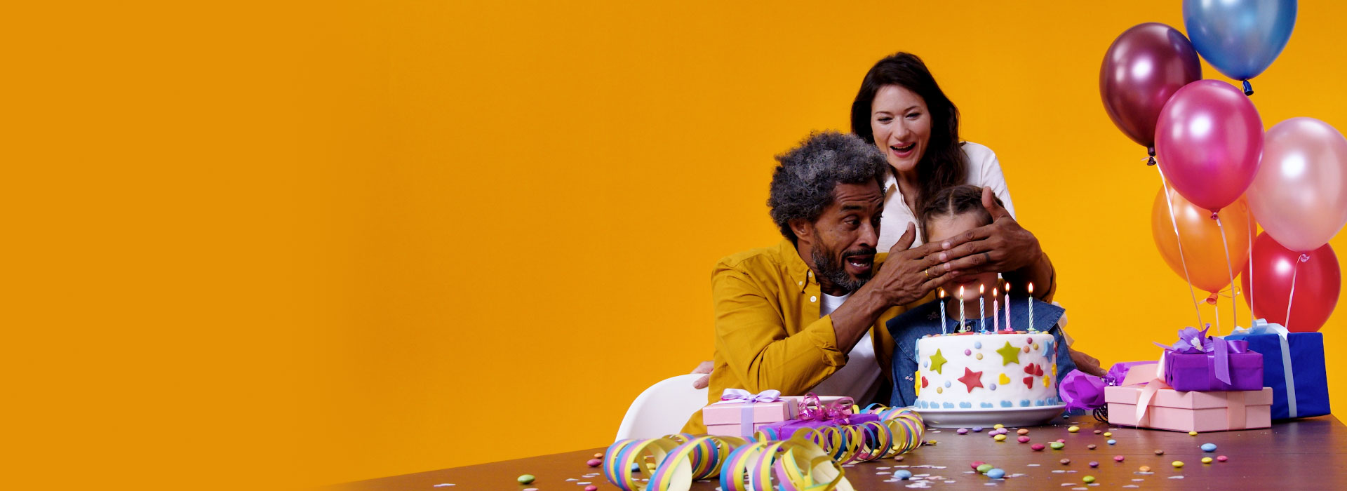Grandfather with Philips HearLink hearing aids and his daughter celebrate his granddaughter's birthday with cake, balloons and gifts.
