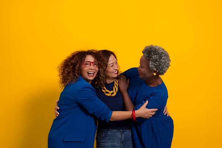 Three middle-aged women of different ethnicities dressed in blue hugging and smiling.  