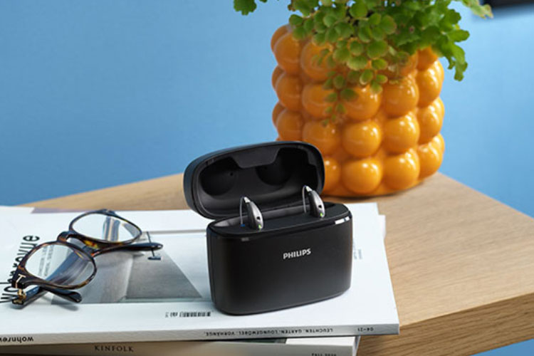Taşınabilir Charger Plus'ın içindeki Philips HearLink şarj edilebilir işitme cihazı, ahşap bir sehpanın üzerinde bir derginin, gözlüğün ve sarı bir saksının yanında duruyor